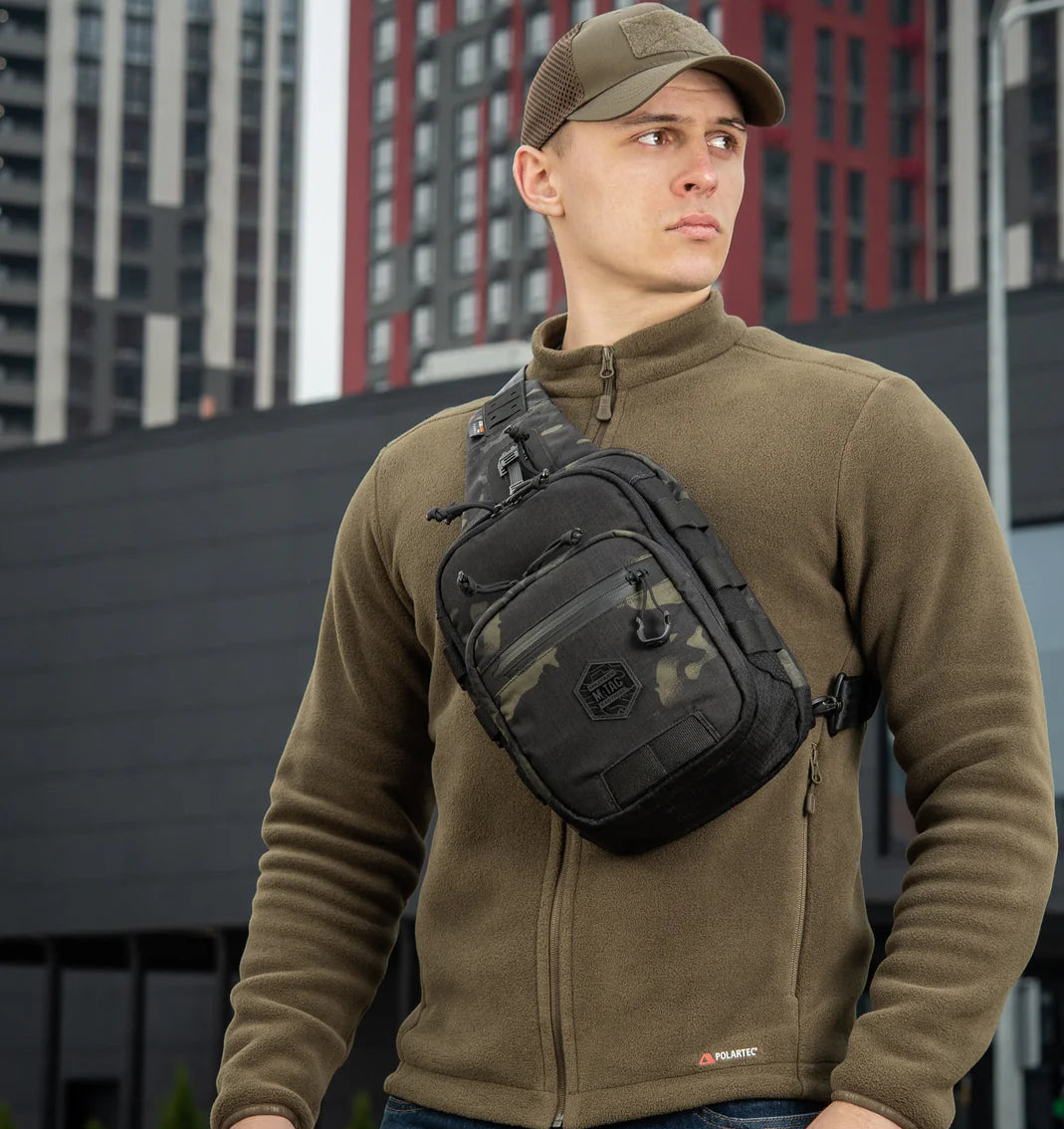 Man wearing a green jacket and cap with a black sling bag in an urban setting